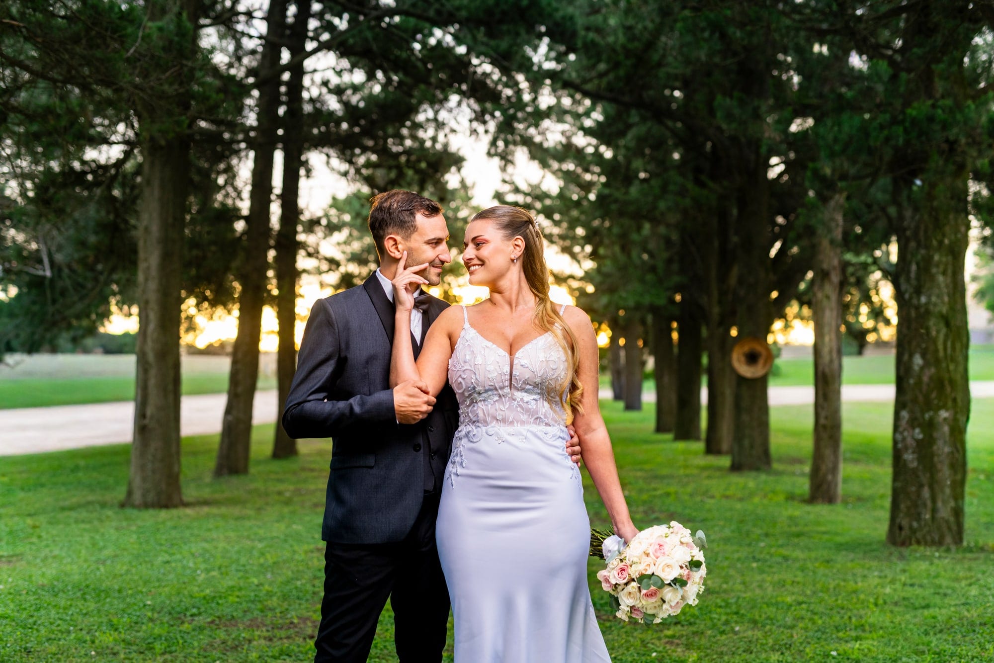 Los novios caminando entre los árboles del parque, con la luz dorada del atardecer y el ramo en la mano