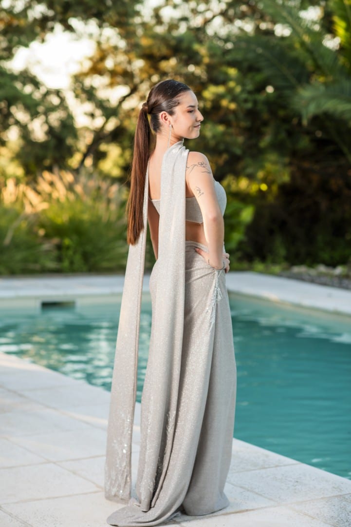 Egresada con vestido largo plateado, de espaldas junto a la pileta, con el jardín encendido por la luz del atardecer