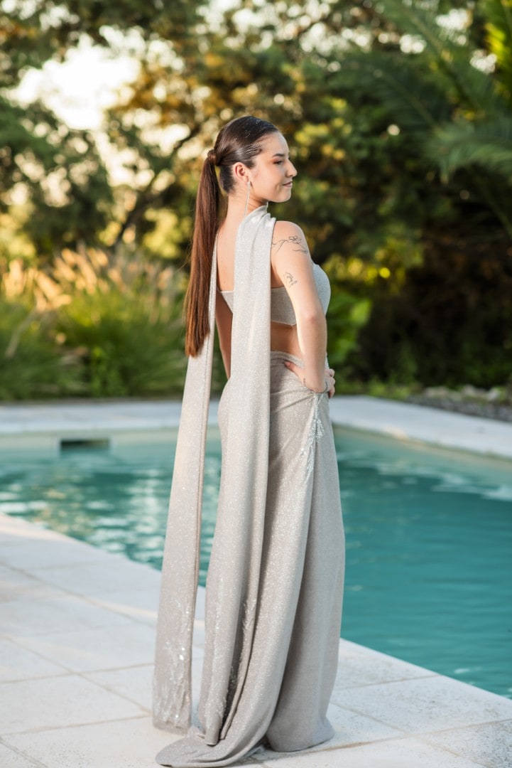 Egresada con vestido largo plateado, de espaldas junto a la pileta, con el jardín encendido por la luz del atardecer