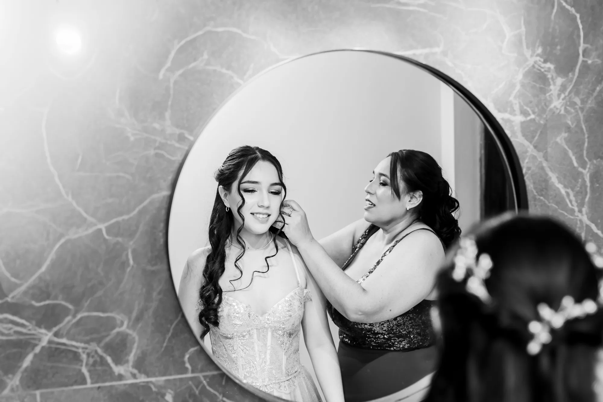 Quinceañera con vestido plateado ajustándose un aro frente a un espejo circular, sobre una pared de mármol azulado